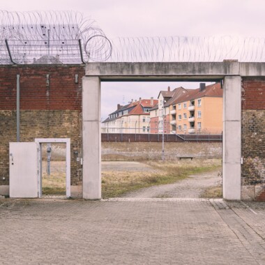 Ein Bild im Innenhof der JVA Rennelberg. Ein großes Tor in einer hohen Backsteinmauer, dahinter das ehemalige Gefängnisgelände. Über der Mauer verläuft Stacheldraht, im Hintergrund sind die umliegenden Wohnhäuser.